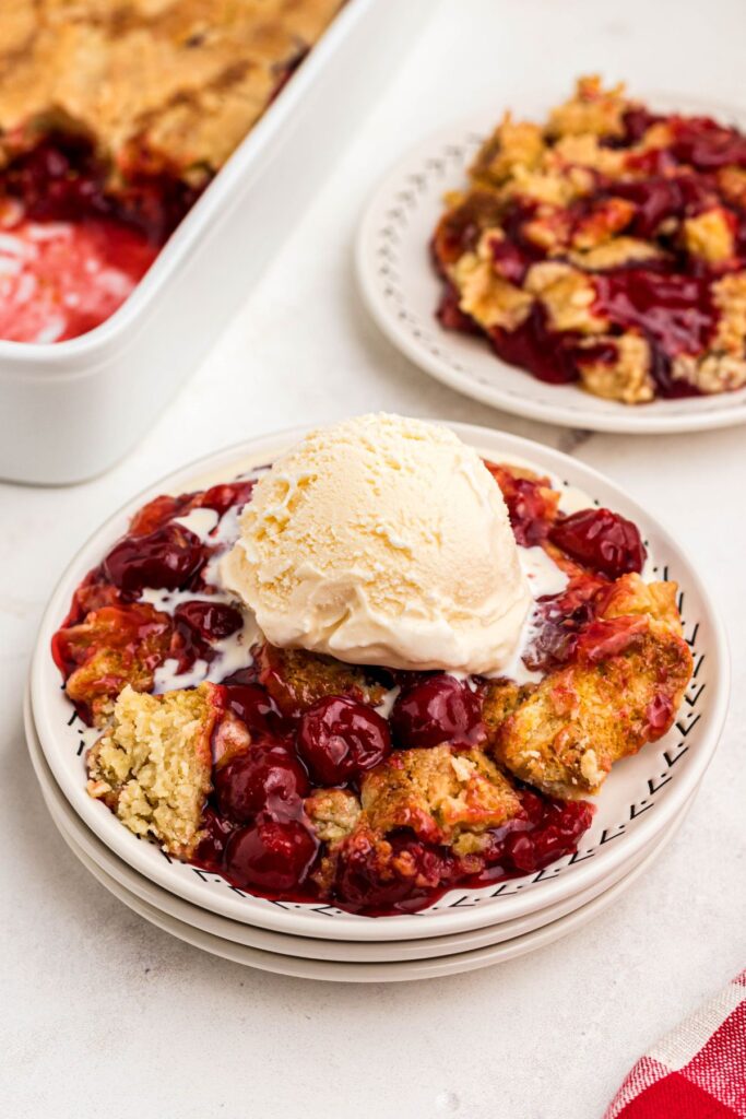 Golden crusted cherry dump cake on white plates topped with vanilla ice cream. 