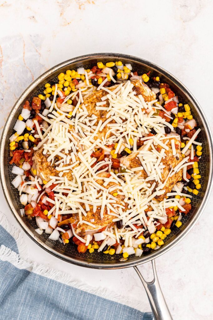 Seasoned seared chicken surrounded by corn, onions, black beans, tomatoes, and shredded cheese, in a large skillet, before cooking. 