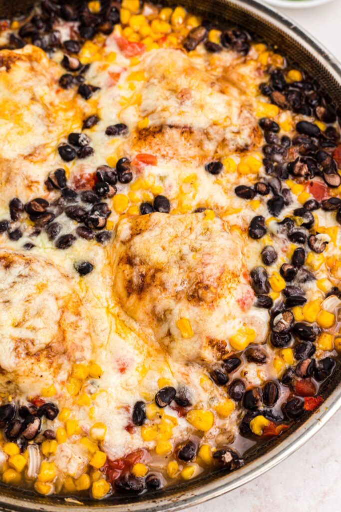 Cheesy chicken in a large skillet, surrounded by corn, black beans, onions, and tomatoes after being cooked. 