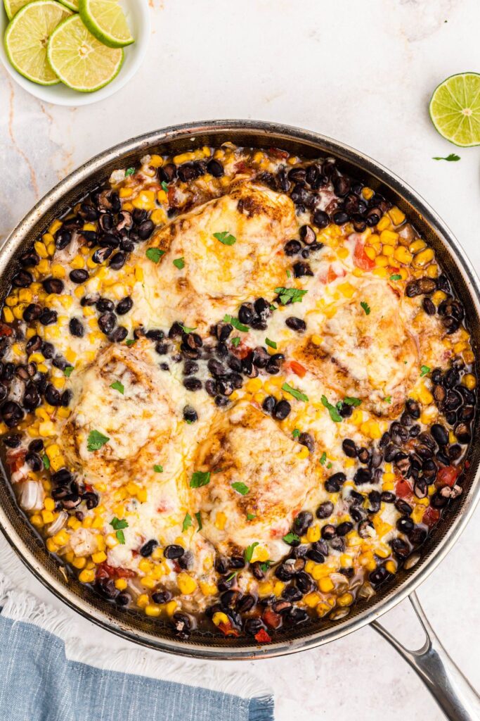 Cheesy seasoned cowboy chicken in a large skillet, surrounded by beans, corns, and tomatoes. 