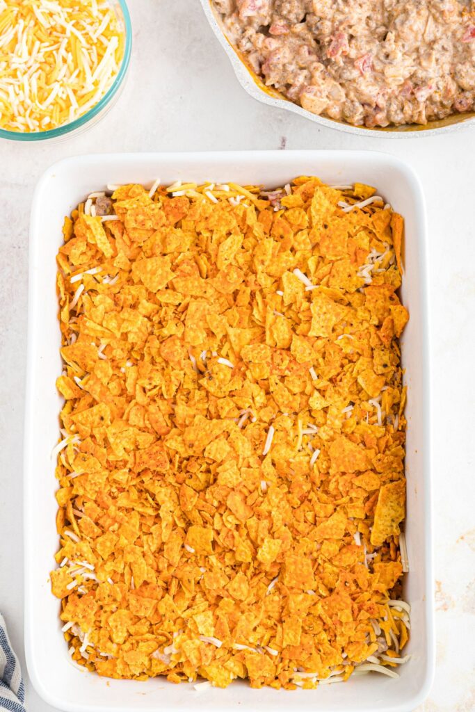 Crushed Dorito's layered with meat mixture and shredded cheese in a white casserole dish. 