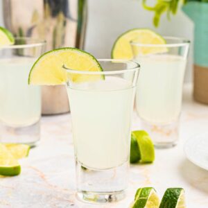 Cold Kamikaze shots in shot glasses with lime wedges and lime curls on a marble table in front of a cocktail shaker.