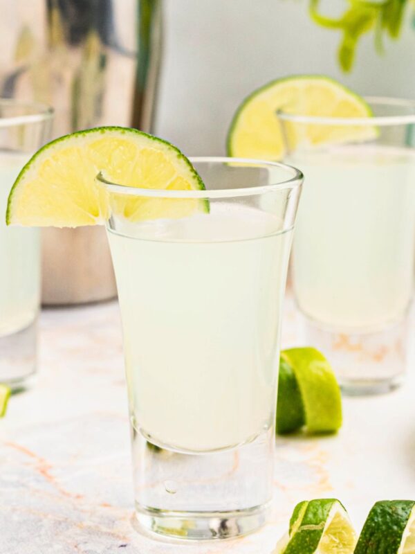 Cold Kamikaze shots in shot glasses with lime wedges and lime curls on a marble table in front of a cocktail shaker.