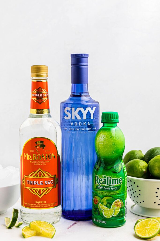 Ingredients needed to make Kamikaze shots on a marble table, along with a bowl of ice, and a bowl of limes. 