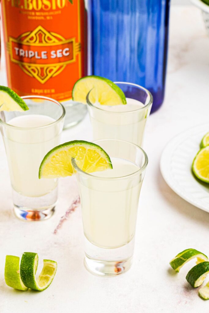 Crisp and refreshing shots in shot glasses with lime wedges and curled limes on a marble table. 