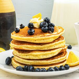 Golden fluffy pancakes stacked on a white plate, topped with blueberries and butter.