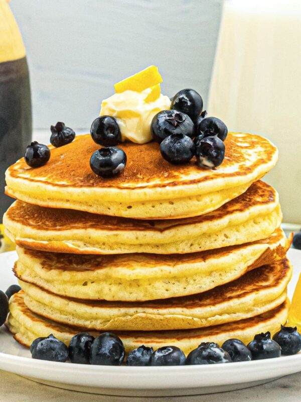 Golden fluffy pancakes stacked on a white plate, topped with blueberries and butter.