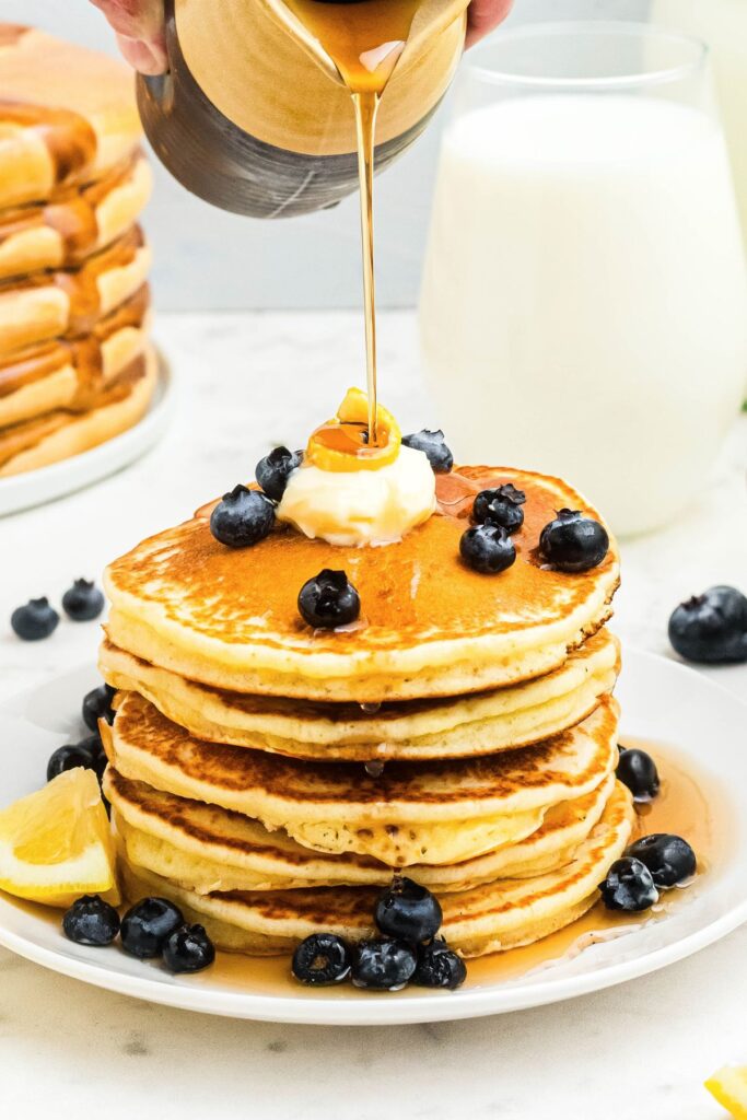 Golden fluffy lemon pancakes stacked on a white plate with syrup being poured on top. 