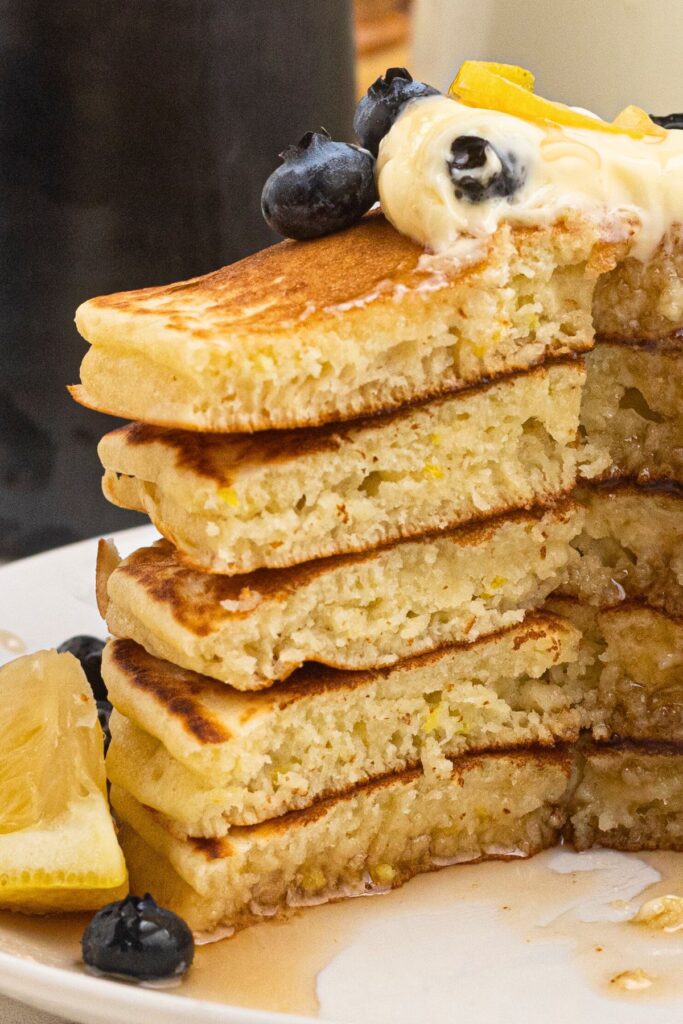 Light and fluffy pancakes stacked on a white plate, drizzled in syrup with butter and blueberries on top.