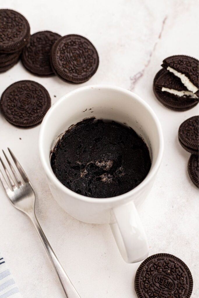 Chocolate cake cooked in a coffee mug, on a marble table with Oreo cookies scattered around the mug. 