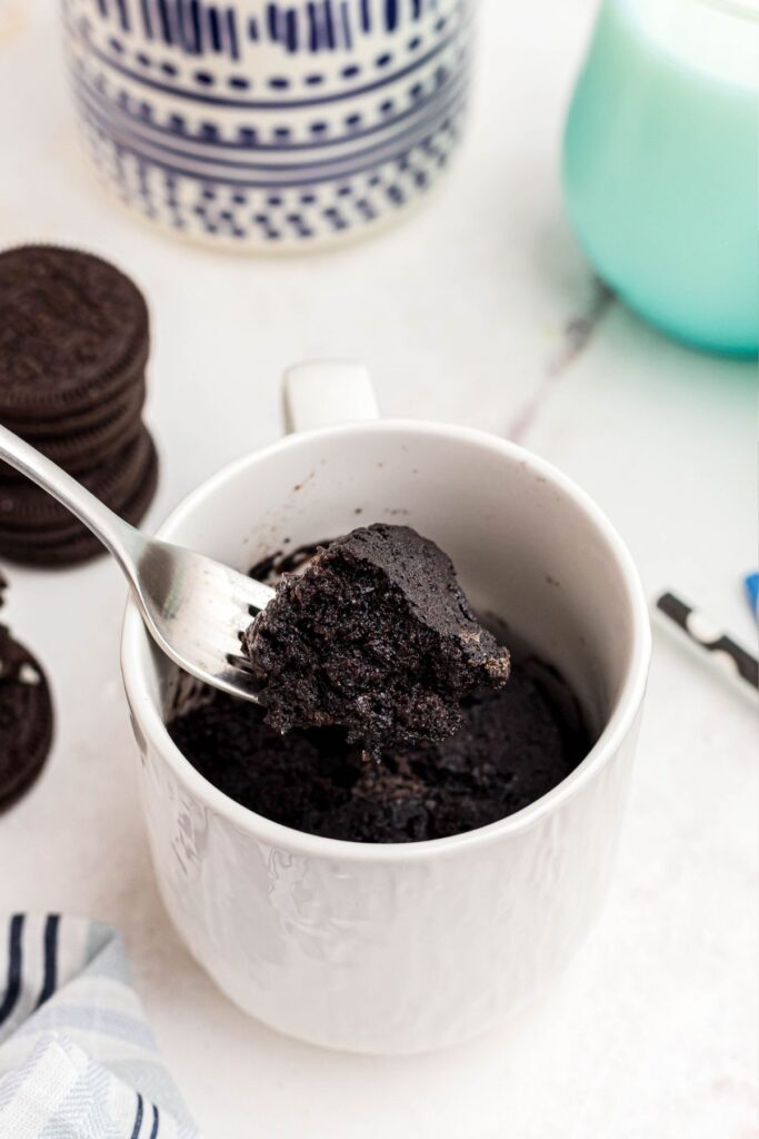 Scoop of chocolate cake being taken out of the mug on a fork with Oreos and glasses of milk on the table. 