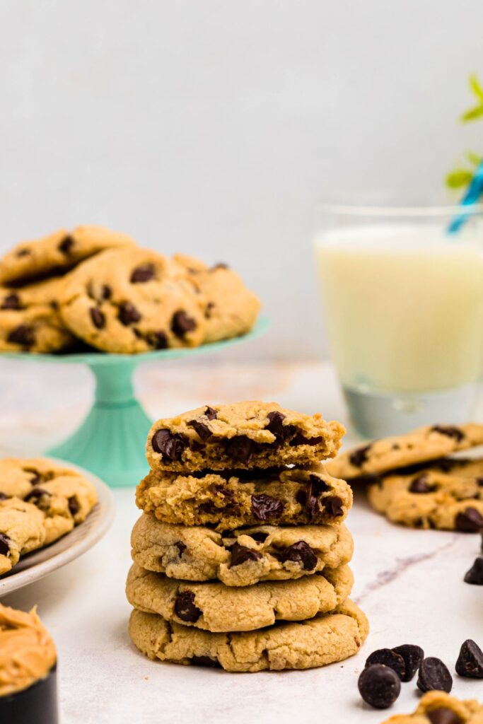 Golden cookies stacked in front of milk and other cookies with the top cookie broken in half showing the chocolate chips inside. 