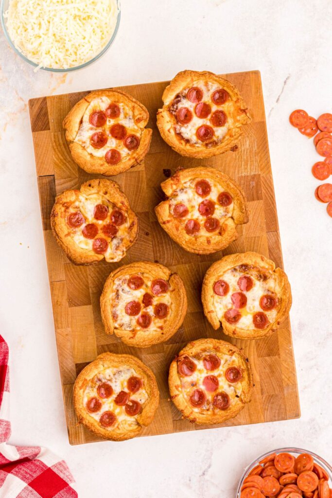 Golden crispy mini pizza cupcakes on a wooden cutting board, with mini pepperonis scattered on a marble table. 