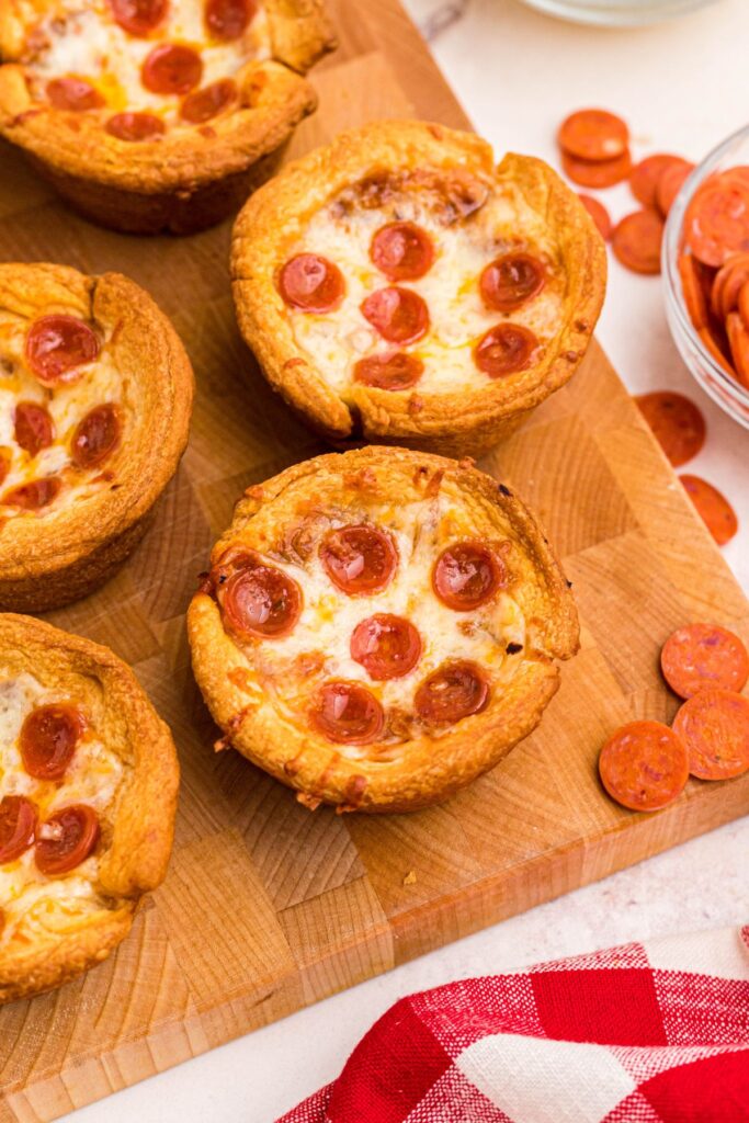 Golden pizza cupcakes on a wooden cutting board with mini pepperonis scattered around it. 