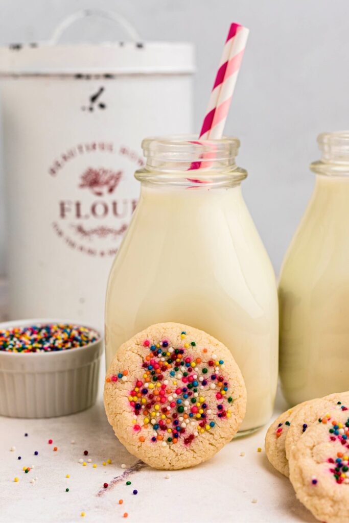 Sugar cookie with sprinkles leaning up against a small bottle of milk with pink straws. 