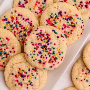 Sugar cookies topped with sprinkles stacked on a rectangle white plate .