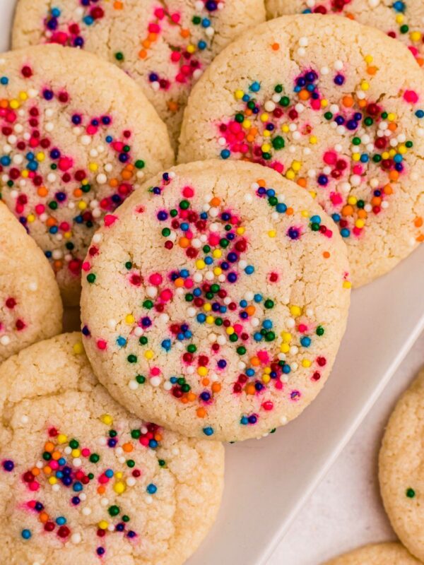 Sugar cookies topped with sprinkles stacked on a rectangle white plate .