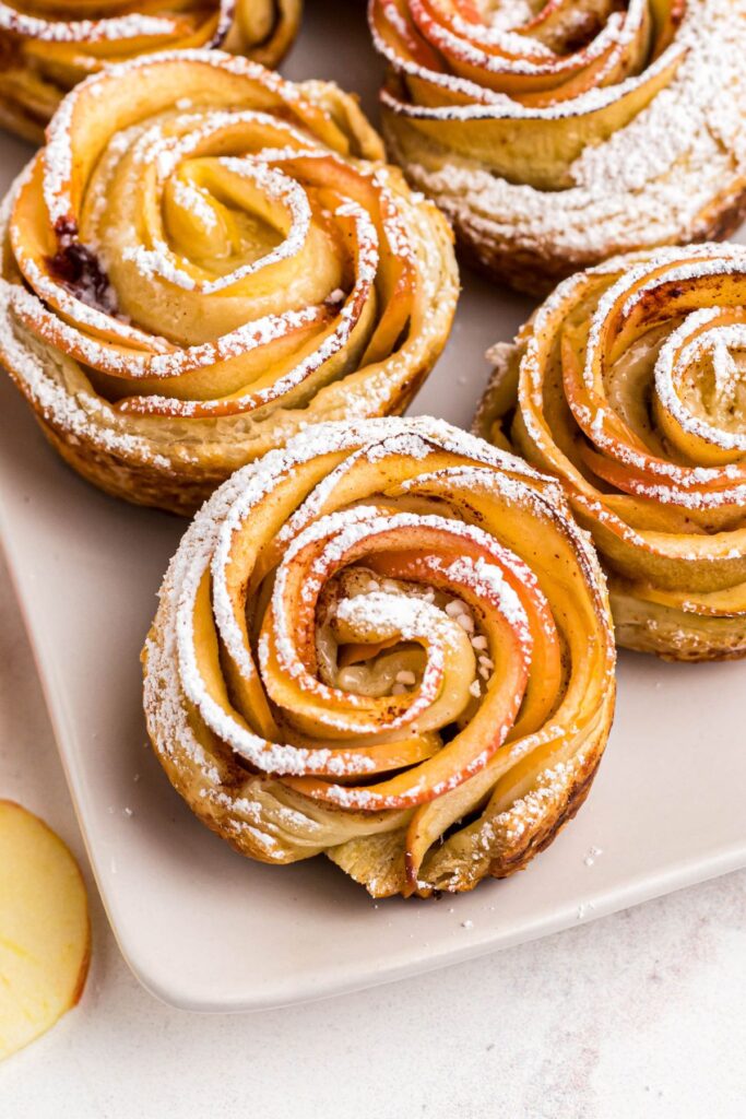 Close up photo of apple rose on a white rectangle plate, then dusted with powdered sugar. 