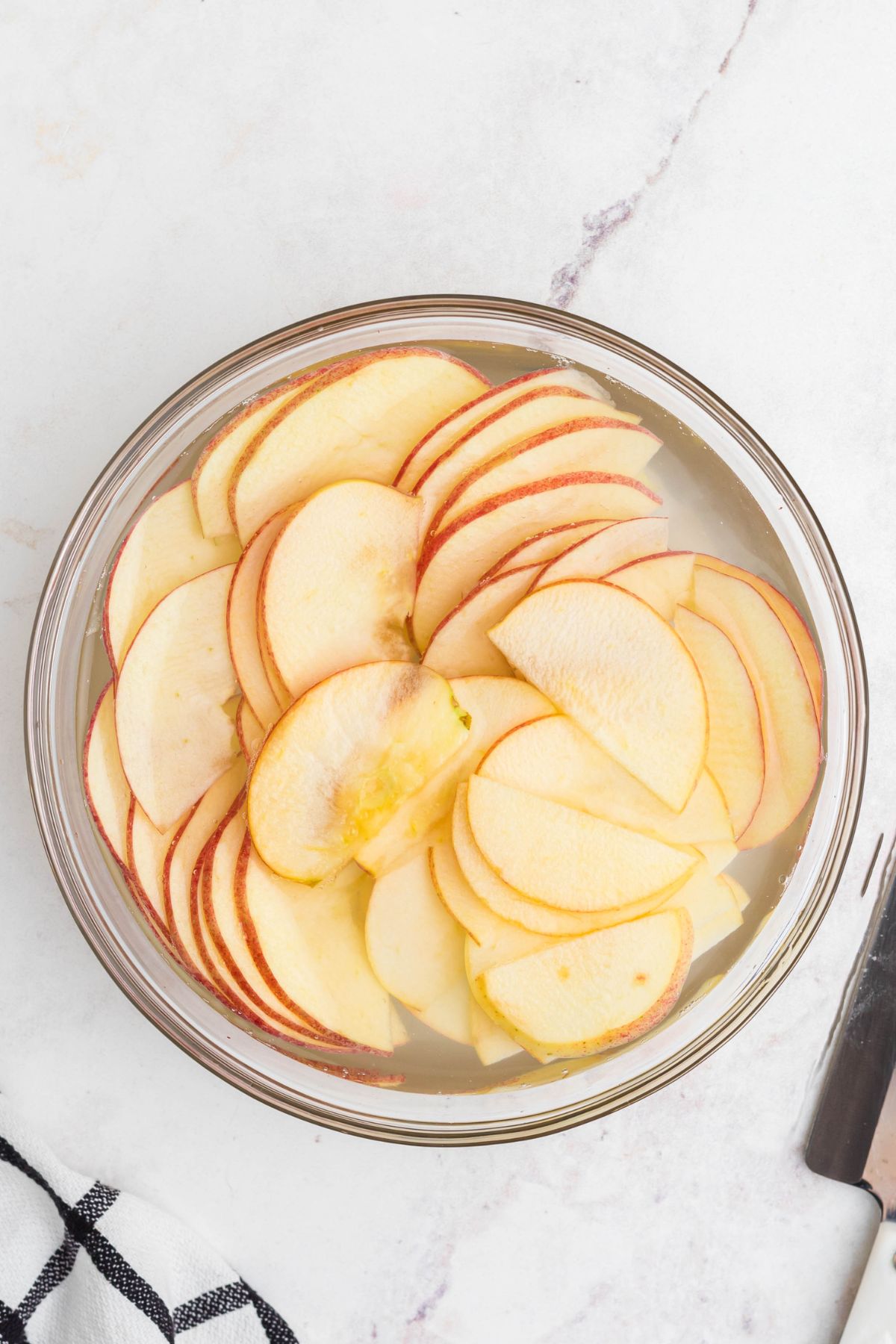 Clear bowl filled with water soaking apple slices, on a marble table. 