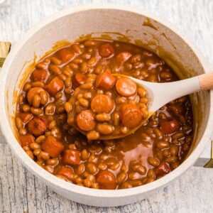 Close up photo of beans and weenies in a saucepan with a large spoon in the center.