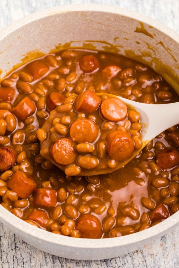 Close up photo of beans and weenies in a saucepan with a large spoon in the center.