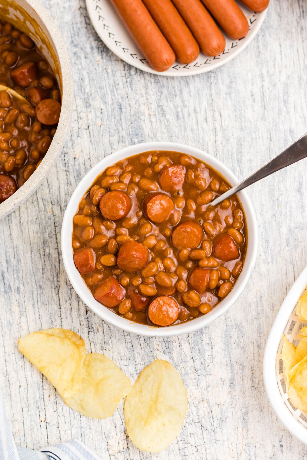 Cut up hot dogs, mixed with beans, in a white bowl with potato chips scattered around the dish. 