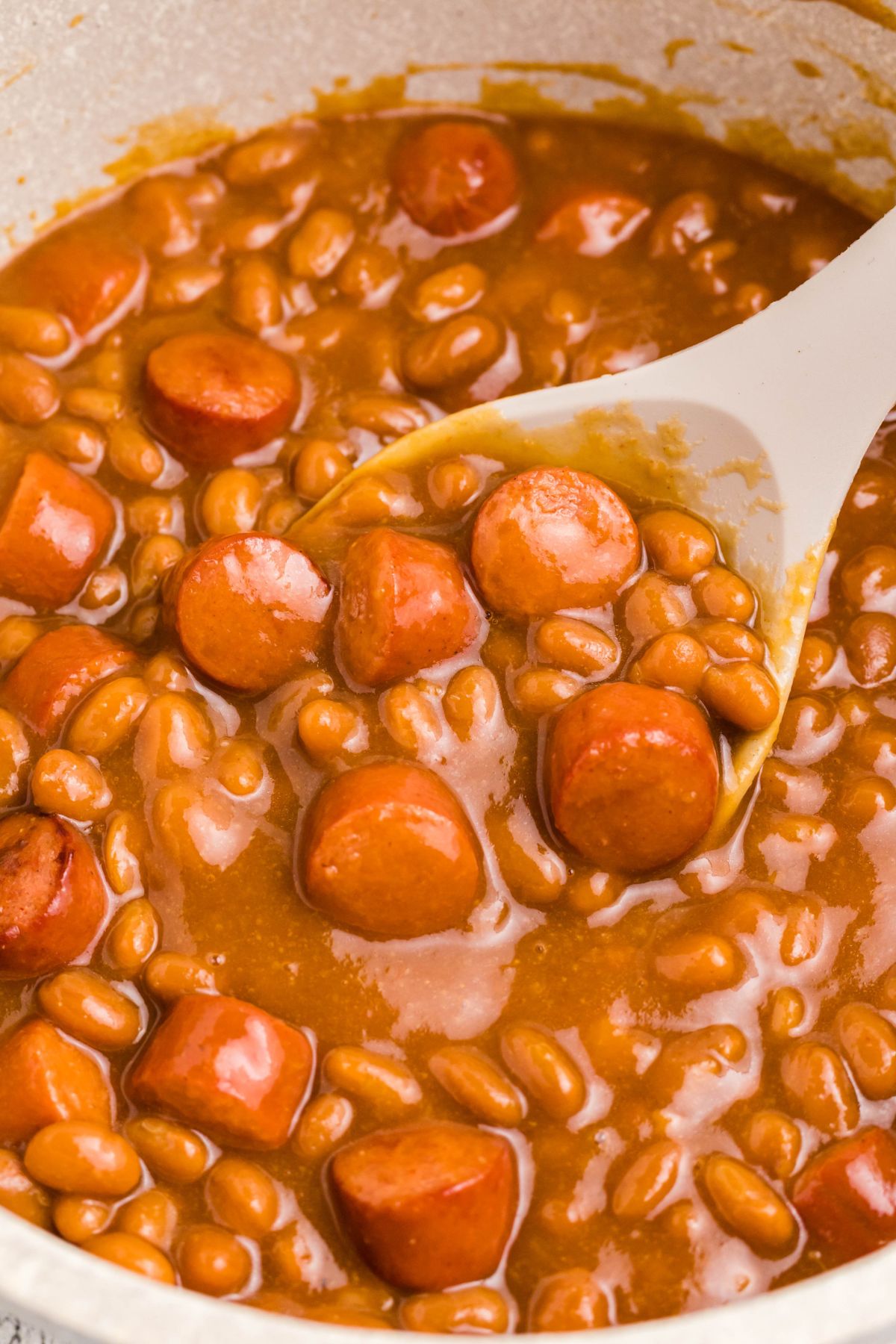 Close up photo of beans and weenies in a saucepan with a large spoon in the center. 