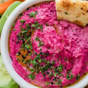 Pink hummus in a white bowl with a cracker in the bowl, garnished with parsley flakes.