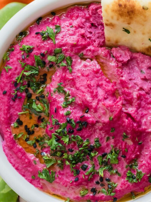 Pink hummus in a white bowl with a cracker in the bowl, garnished with parsley flakes.