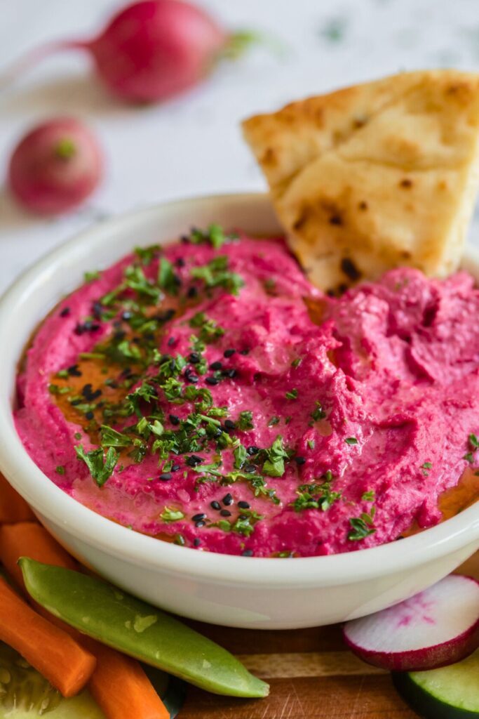 Pink beet hummus in a small white bowl surrounded by vegetables for dipping. 