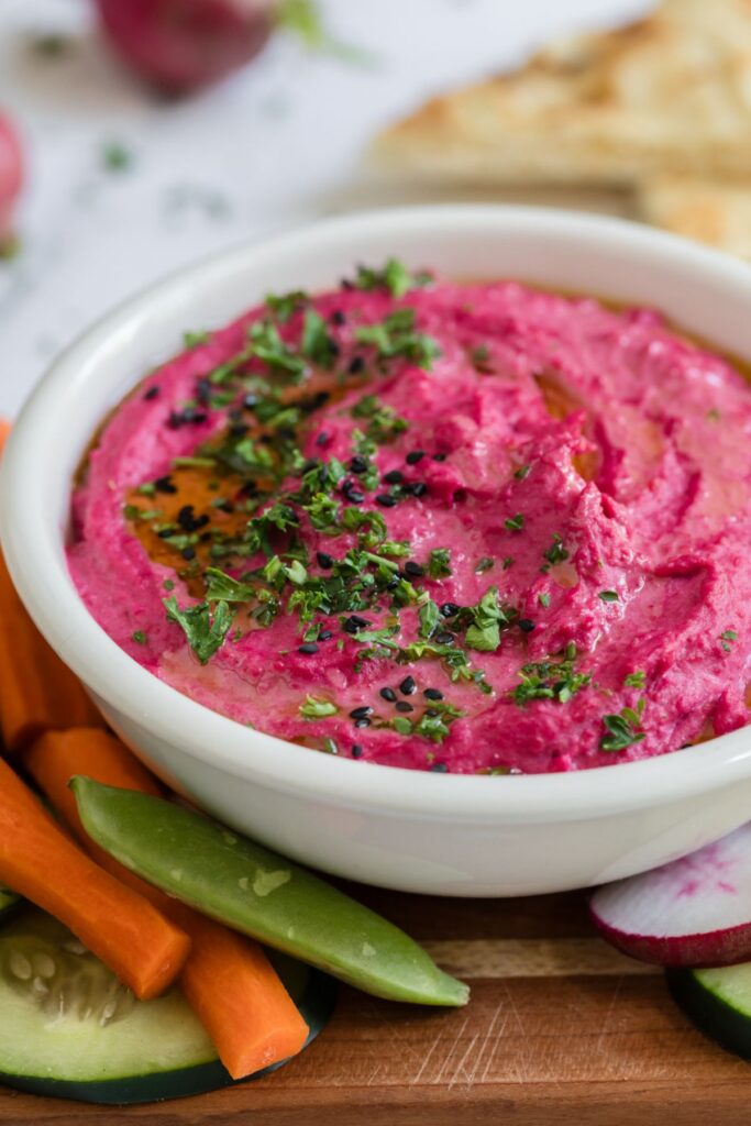 Bright pink hummus in a small white bowl surrounded by veggies. 