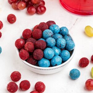 Red and blue candy grapes stacked in a white bowl with grapes scattered on a marble table.