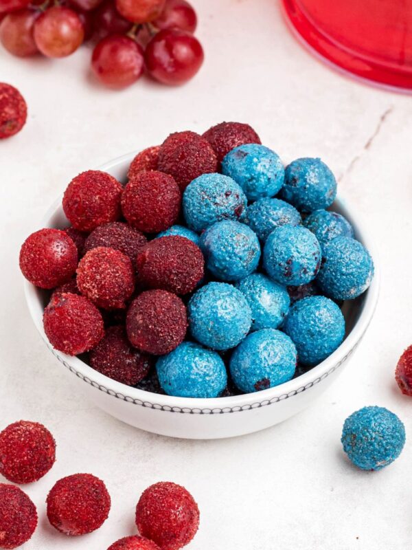 Red and blue candy grapes stacked in a white bowl with grapes scattered on a marble table.