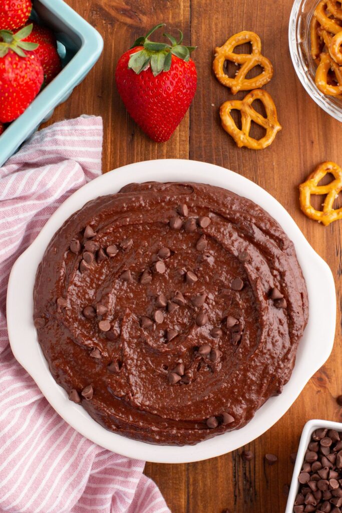 Chocolate hummus in a white serving dish, topped with mini chocolate chips, surrounded by strawberries and pretzels for dipping. 