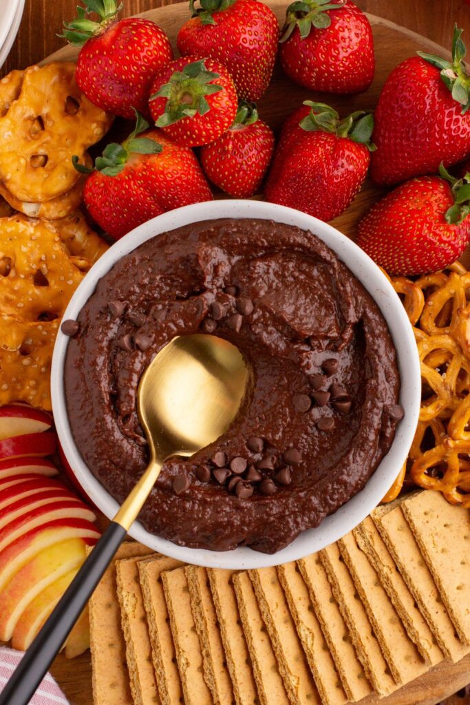 Chocolate hummus in a white bowl with a golden serving spoon in the center, with dipping items like strawberries and pretzels around the bowl. 