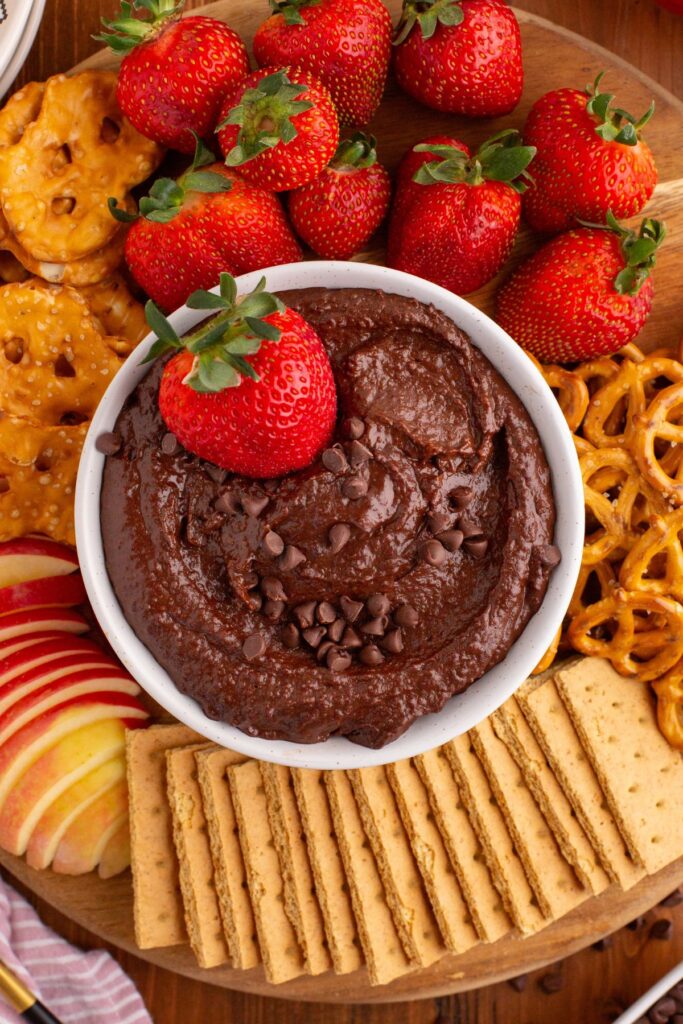 Smooth and creamy chocolate hummus in a white bowl with crackers and fruit on a serving platter. 