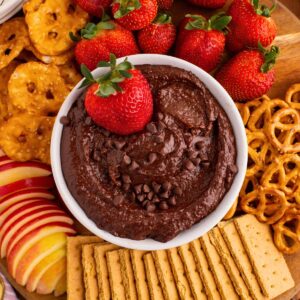 Chocolate hummus in a white bowl with a golden serving spoon in the center, with dipping items like strawberries and pretzels around the bowl.