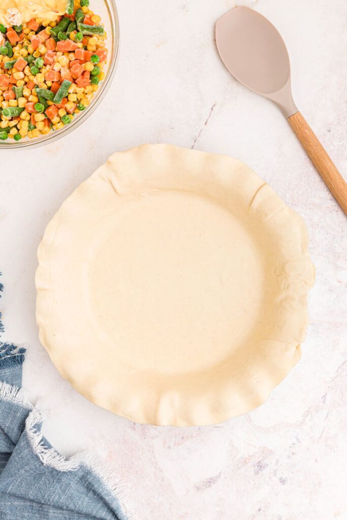 Rolled out pie crust in a pie plate with a bowl of veggies and other filling ingredients on a marble table. 