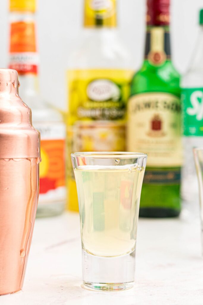 Green tea shot in a small shote glass next to a gold cocktail shaker. 