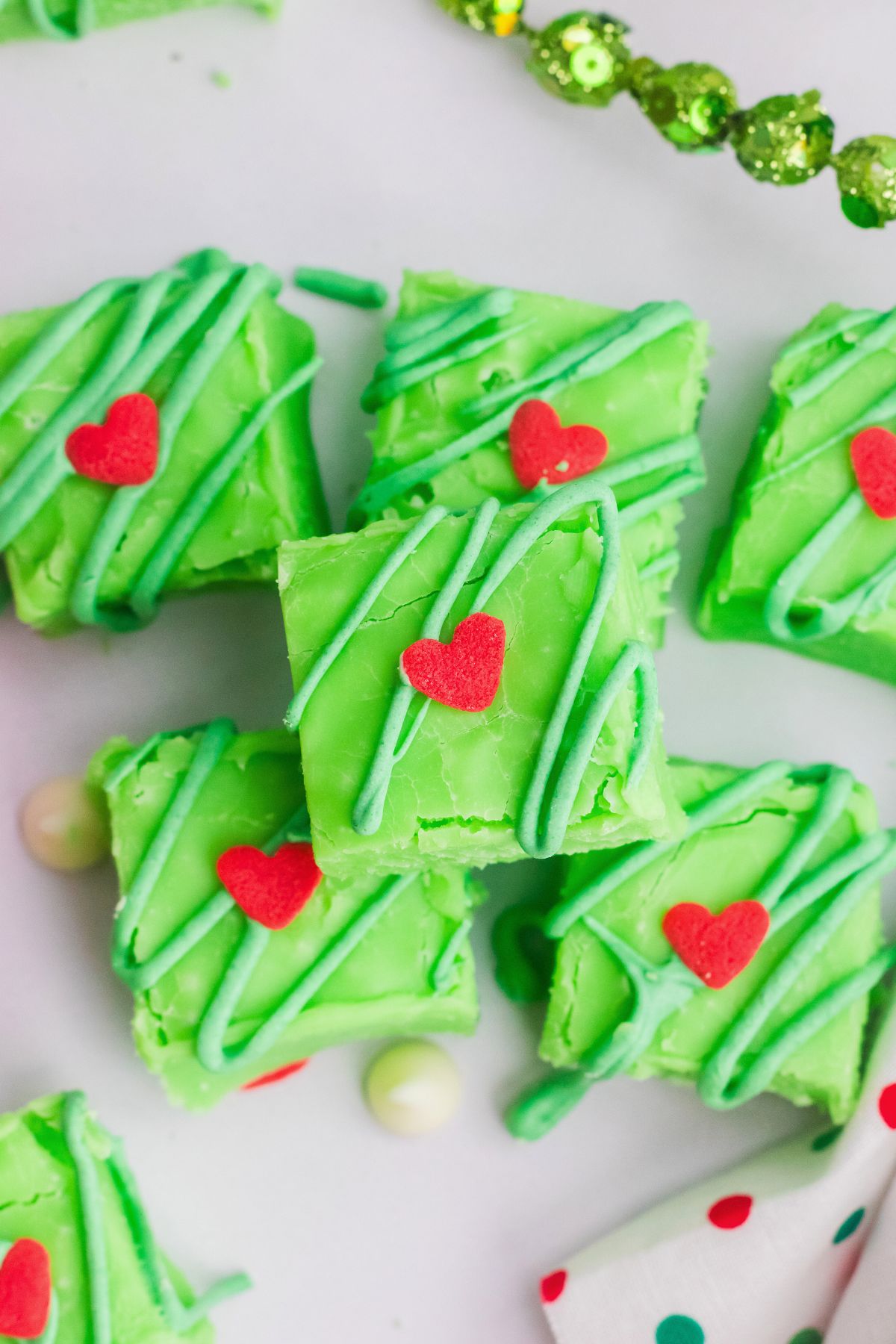 Square green fudge pieces topped with small red hearts, stacked on a white table. 