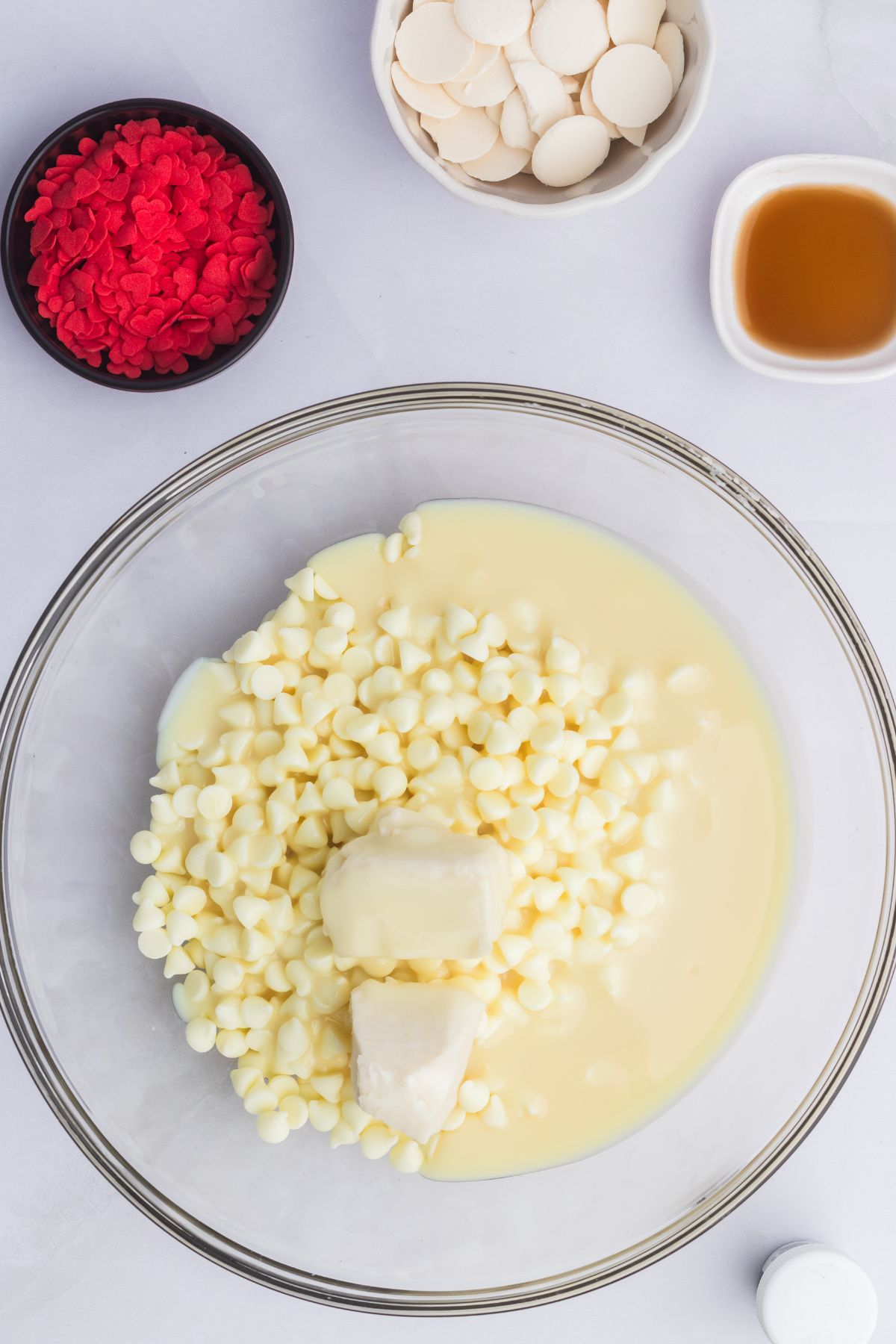 White chocolate chips being melted in a clear glass bowl with other ingredients on the table. 