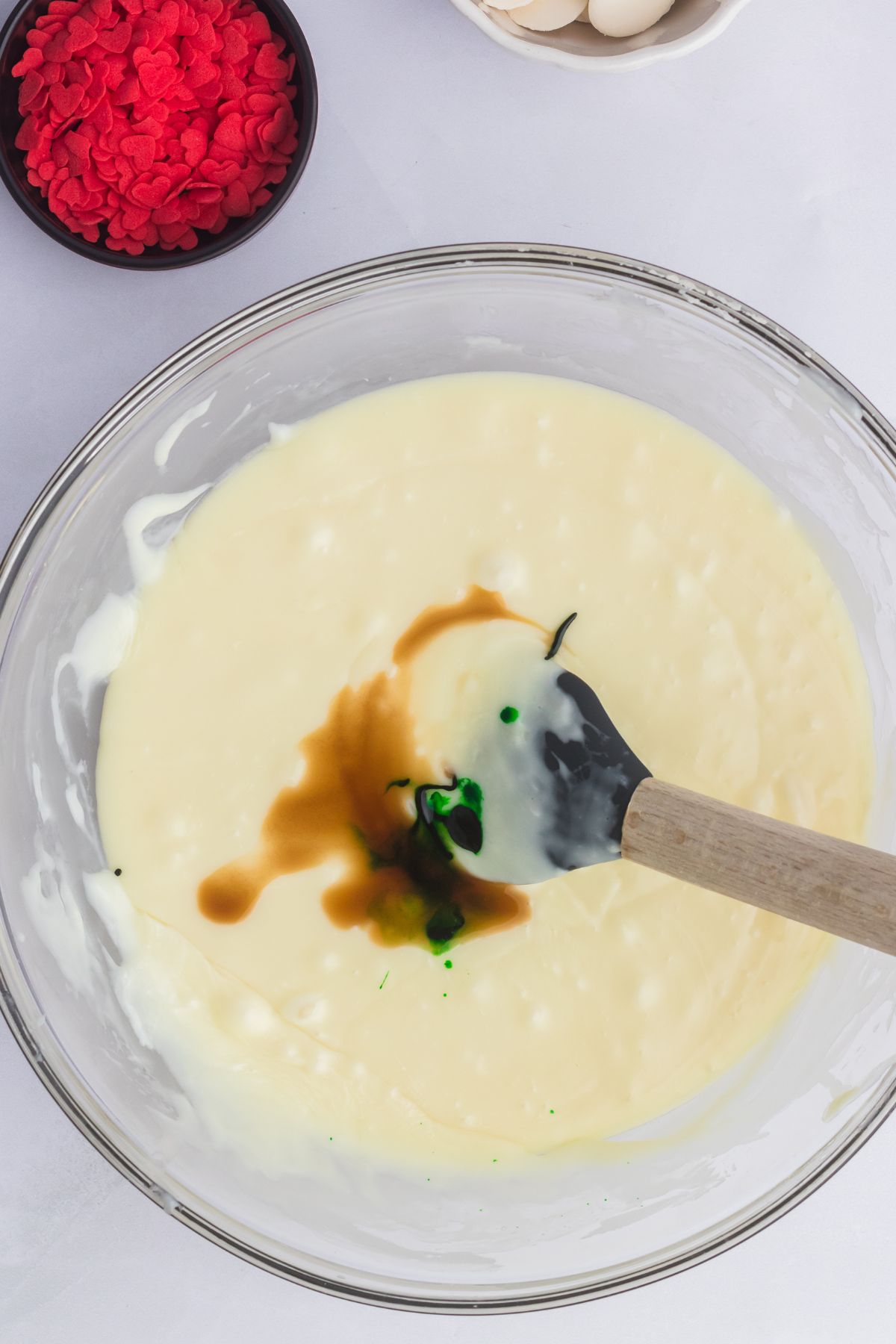 Melted white chocolate with green food coloring being added to it in a large clear glass bowl. 