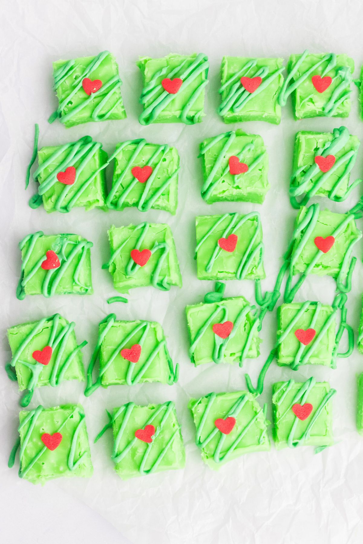 Bright green Grinch fudge, cut into squares on parchment paper with small red hearts added to the tops of each piece. 