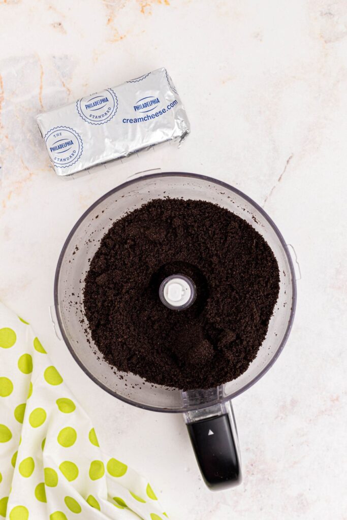 Crumbled Oreos in a food processor bowl with a package of cream cheese on a marble table. 