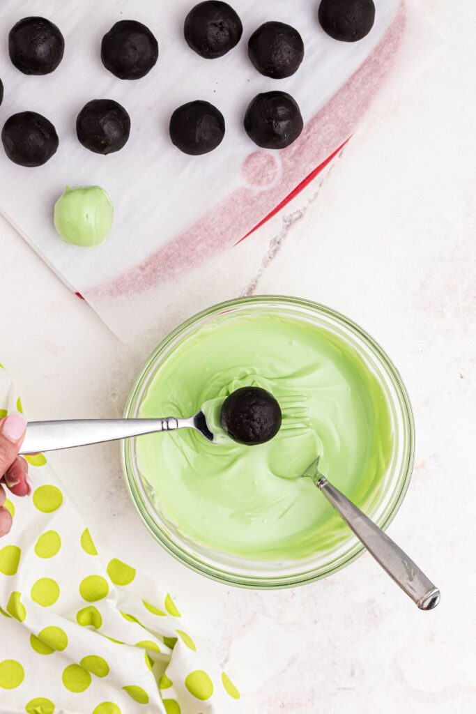 Oreo ball being dipped into melted green candy melts with remaining balls on parchment.