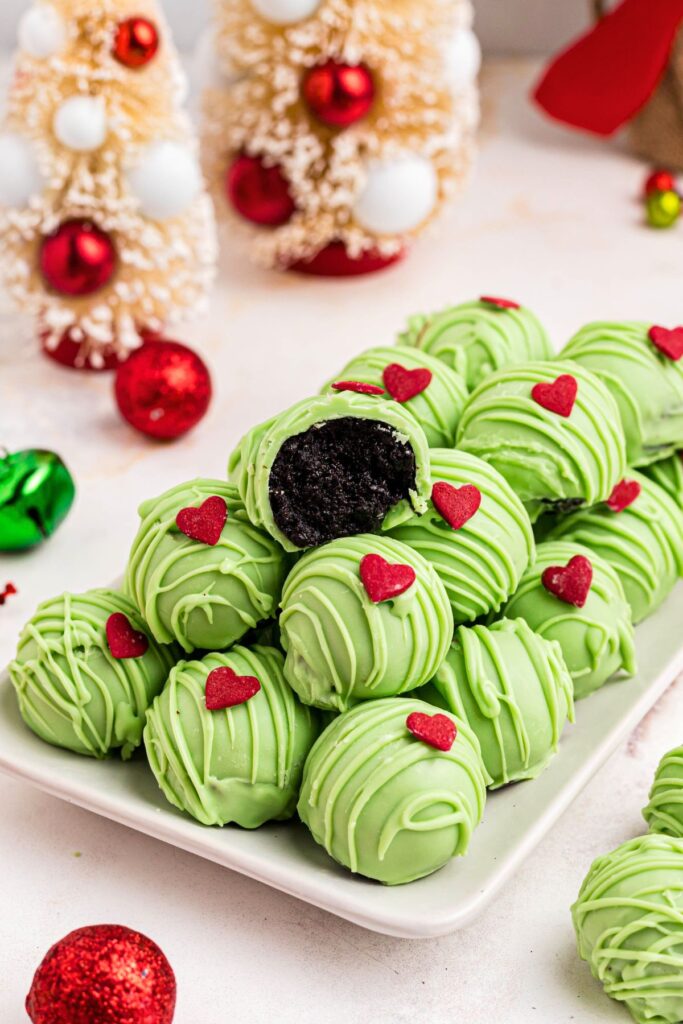 Green Grinch Oreo balls stacked on a white plate with one on top with a bite out and ornaments on the table surrounding the bites. 