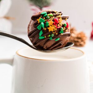 White mug of hot milk with a spoon holding a chocolate cocoa bomb over the milk.