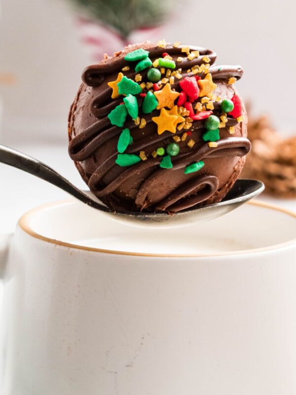 White mug of hot milk with a spoon holding a chocolate cocoa bomb over the milk.