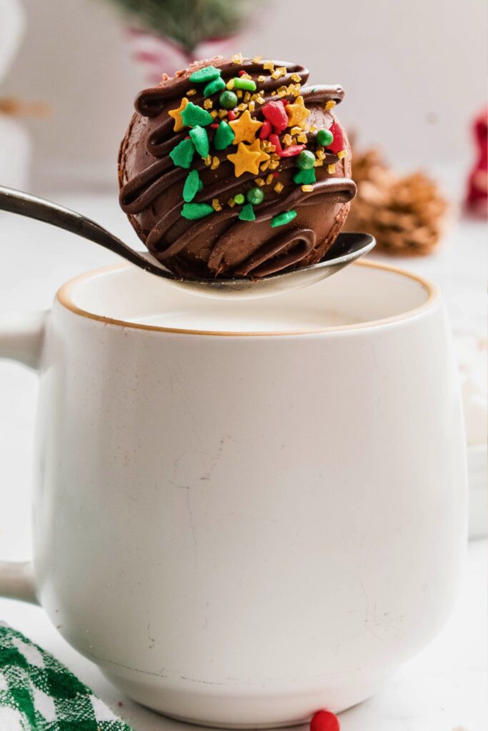 White mug of hot milk with a spoon holding a chocolate cocoa bomb over the milk.