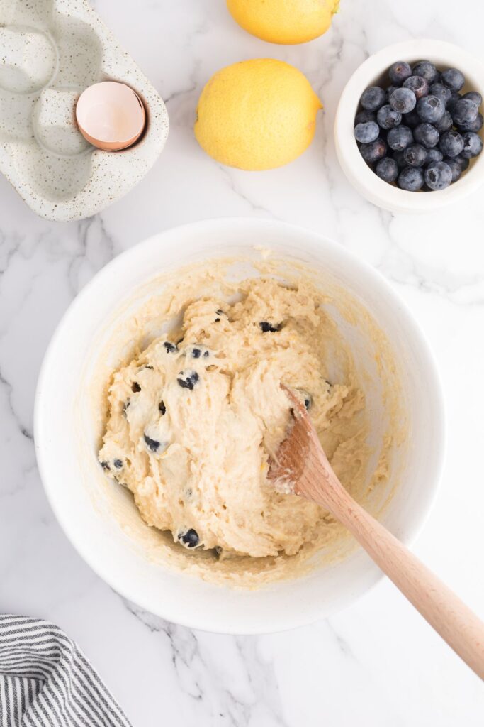 Blueberries mixed into batter in a white bowl with a wooden spoon. 